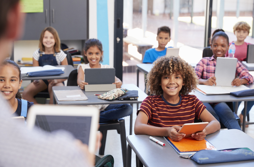 Students in a classroom listening to their teacher while working on computer science on tablets.