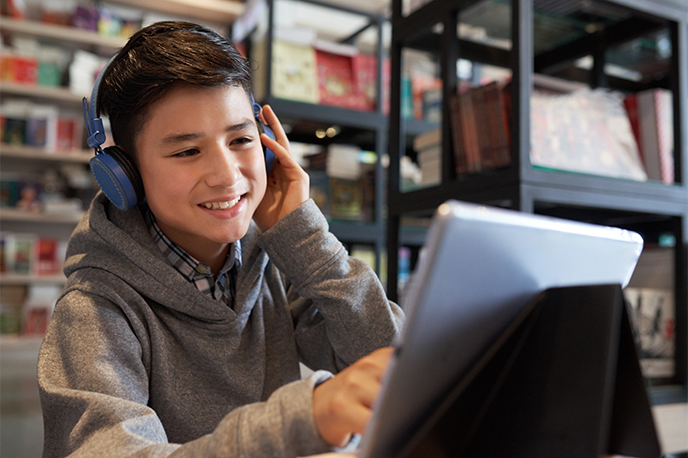 A smiling student wearing headphones and pointing at a tablet while sitting in a library