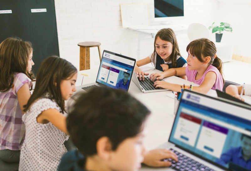 Two middle school students working on a laptop together with code shown on a display behind them.