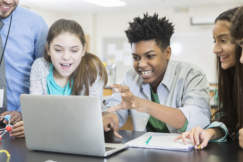 Group of high school students and an instructor gathered around a laptop within a science classroom