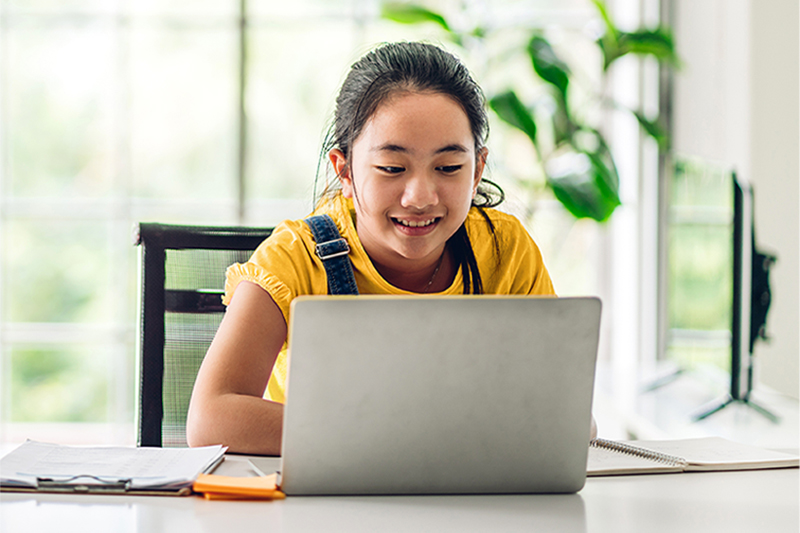 Elementary school student sitting at a table while smiling and typing on a laptop