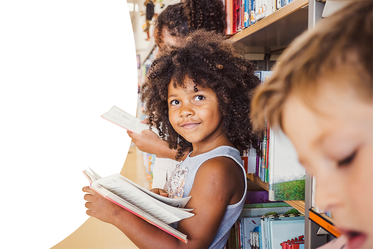 Three elementary students in a library, leaning against a wall of books and reading books.