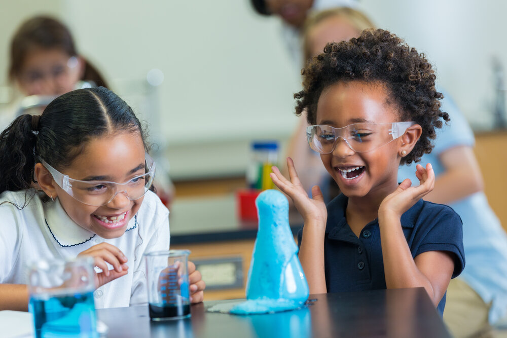 Two young students reacting to a flask overflowing with bubbles in a classroom