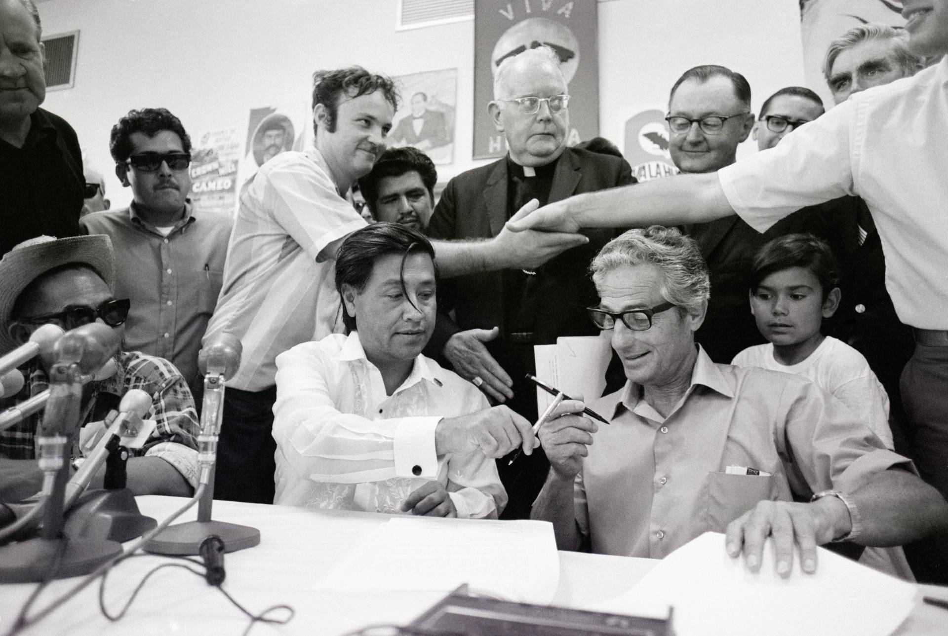 Historical photograph of Ceasar Chavez and John Giumarra sitting at a table exchanging pens, with papers on the table in front of them.